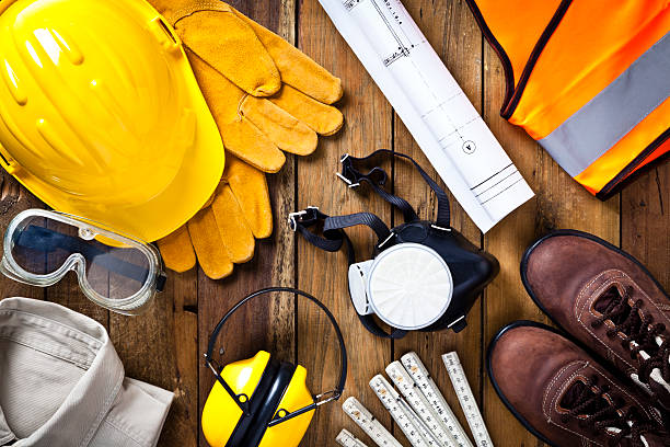 Personal protective workwear and blueprint shot directly from above on rustic wood background. The protective workwear includes hard hat, gloves, earmuff, goggles, steel toe shoes, and safety vest. Predominant colors: yellow and brown. DSRL studio photo taken with Canon EOS 5D Mk II and EF 100mm f/2.8L Macro IS USM